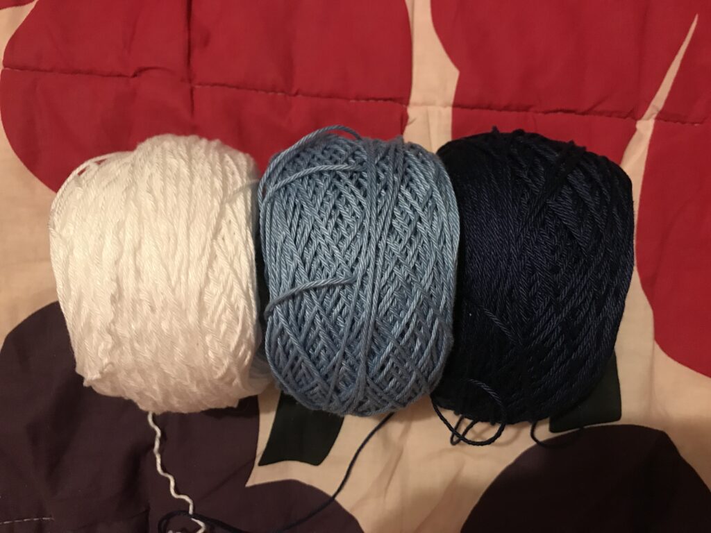 Three skeins of yarn in white, light blue, and navy, on top of a patterned fabric.